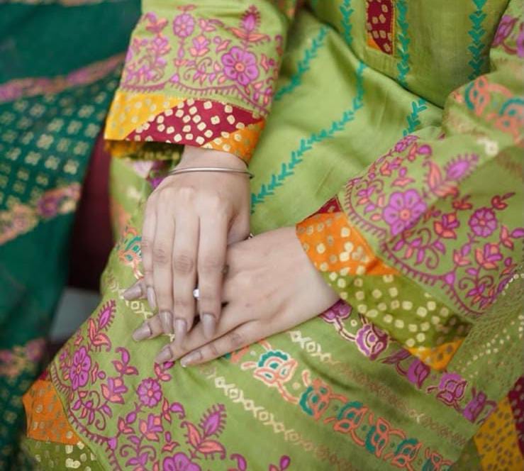 A close-up of a person wearing a bright lime green block printed traditional outfit with colorful floral patterns in pink, orange, yellow, and teal. The person's hands are folded on their lap, showing manicured nails and a thin silver bangle.