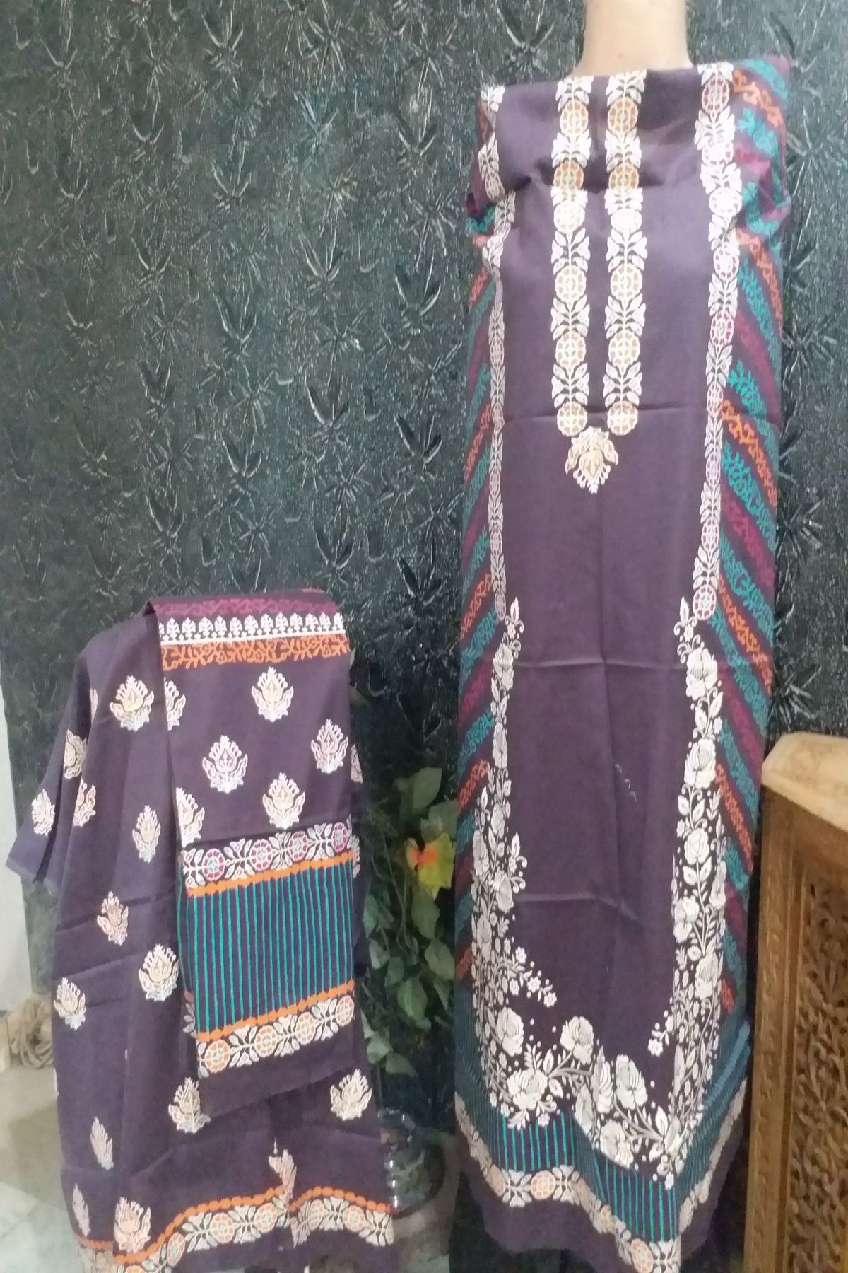 A purple ethnic wear fabric set with white floral prints, teal stripes, and orange border details, displayed on a mannequin and folded fabric piece against a dark textured wall.