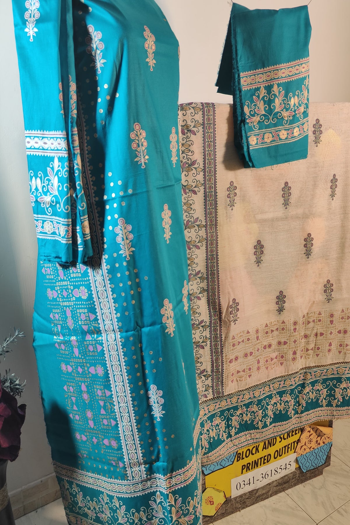 A 3-piece unstitched hand block print suit featuring a vibrant teal fine lawn shirt and trouser with elegant white, pink, and gold floral motifs alongside a contrasting beige and teal chiffon dupatta.