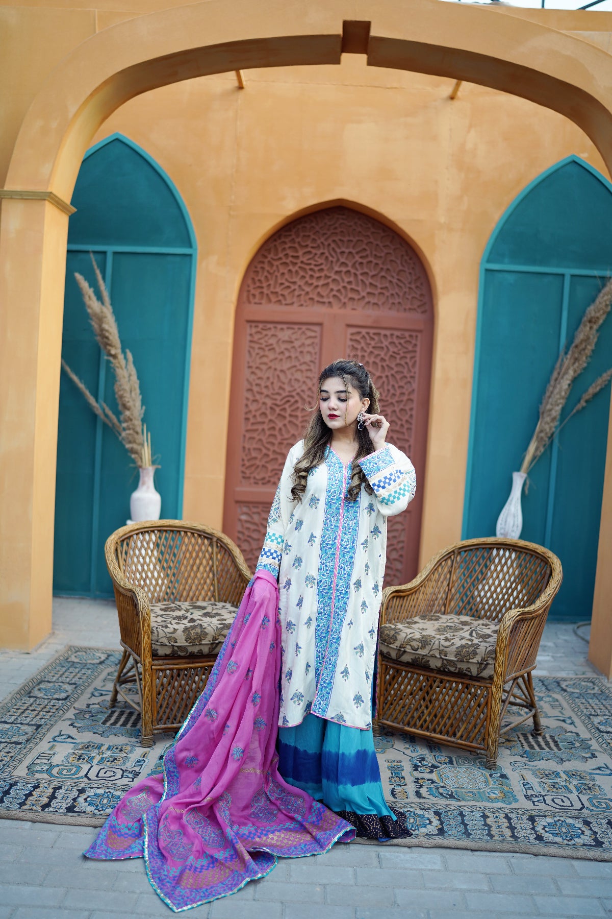 A woman models a traditional South Asian outfit consisting of a white kurta with blue and green embroidery, a blue and green flared skirt, and a vibrant pink dupatta with purple and blue borders, set against a warm ochre-colored archway background with wicker chairs.