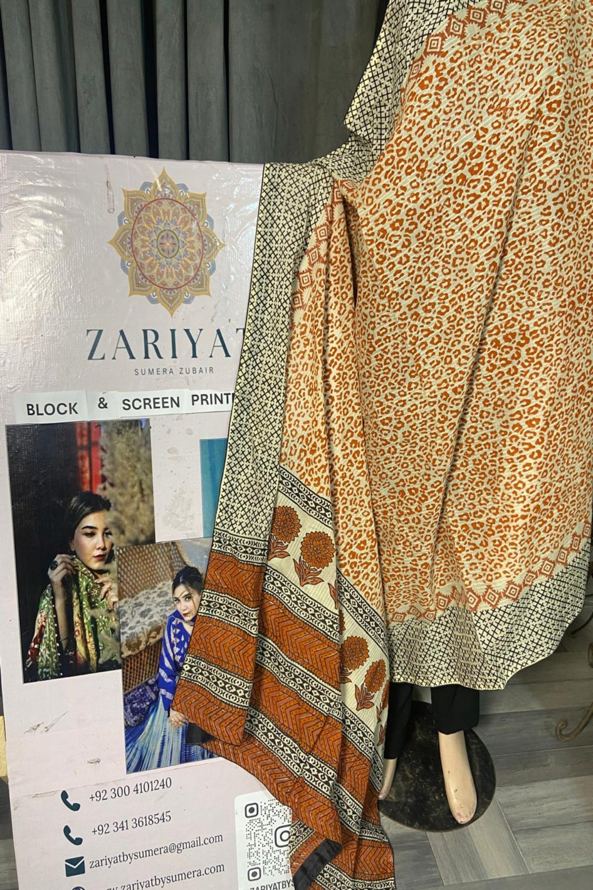 A block printed fabric garment or scarf displayed on a mannequin, featuring an orange and beige leopard-like pattern with a decorative border, alongside a branded paper card for Zariya Sumaira Zuberi.