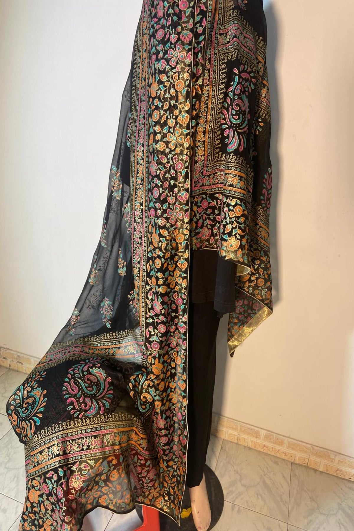 A black block printed traditional outfit draped on a mannequin, featuring intricate multicolor floral and paisley patterns with gold zari detailing, set against a plain indoor wall.