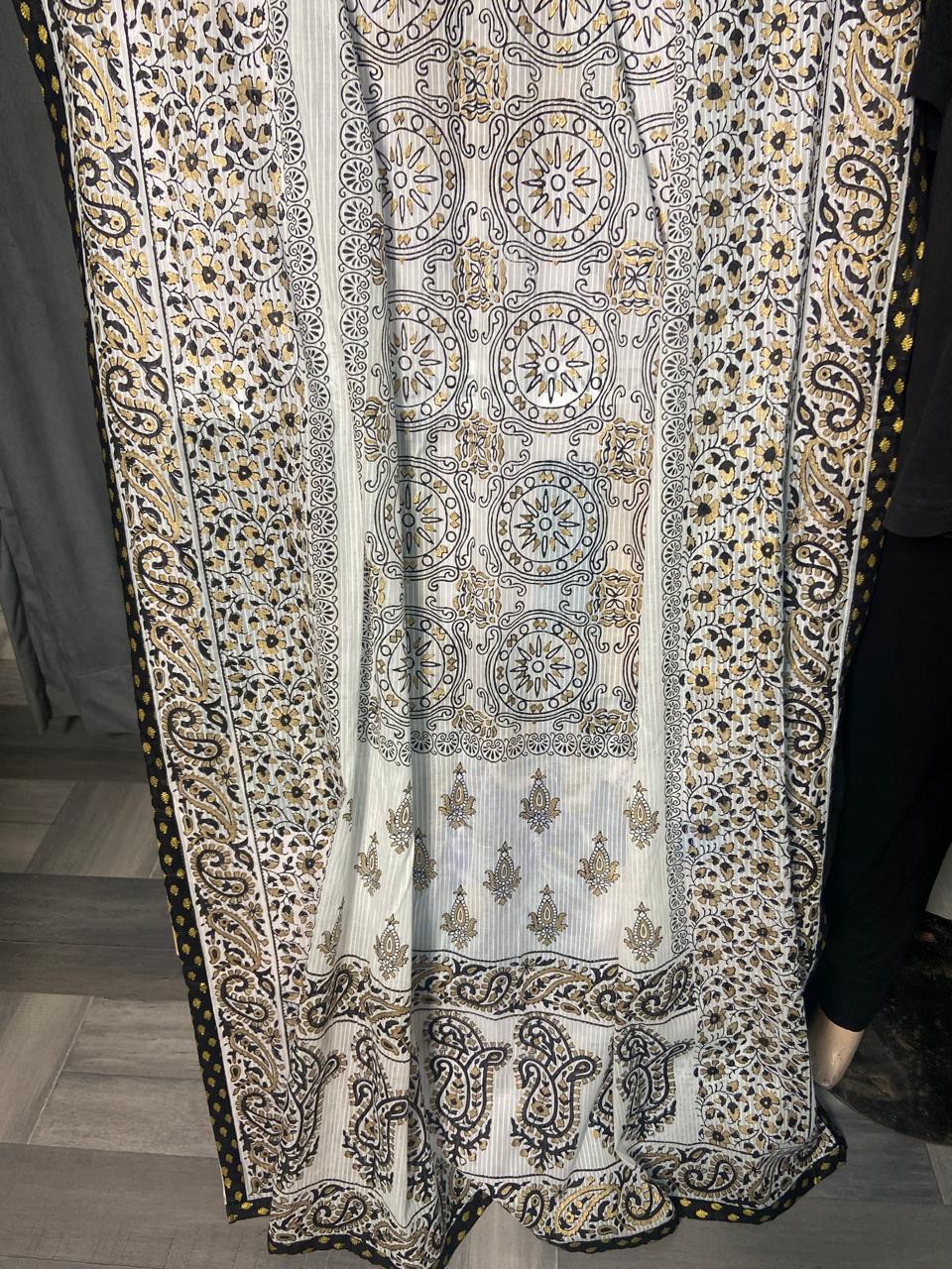 A white and beige block  printed fabric panel with intricate black, gray, and gold geometric and paisley motifs, displayed on a mannequin or hanger against an indoor background.