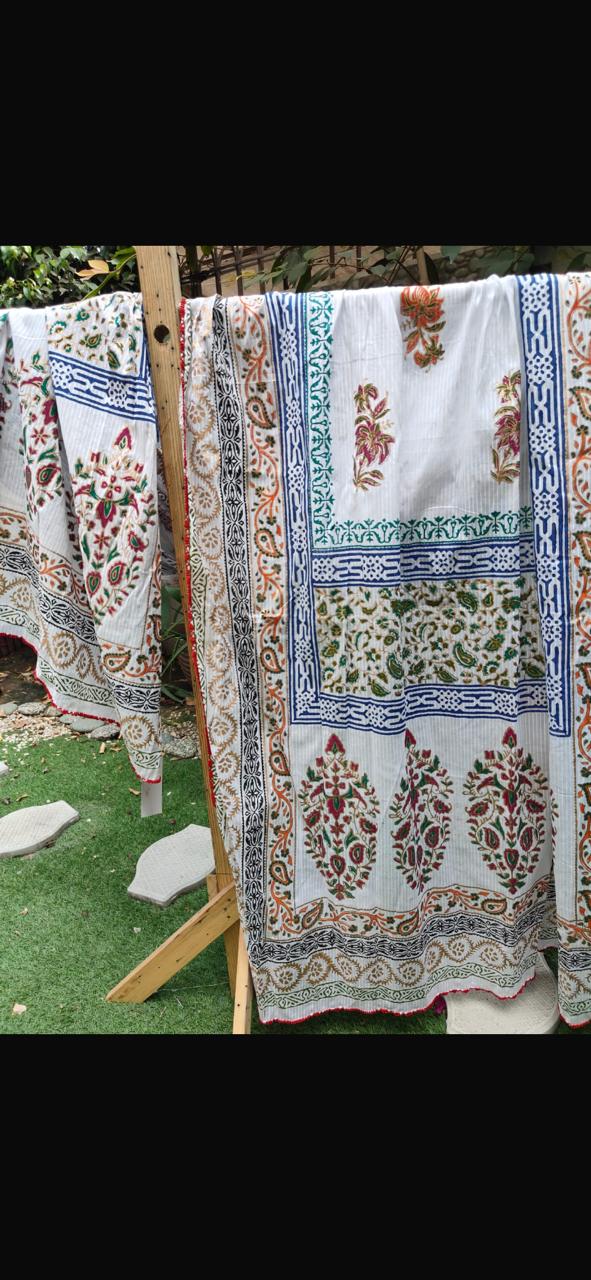 A white cotton fabric with intricate multicolor floral and geometric block prints hangs on a wooden clothesline outdoors on a green lawn.