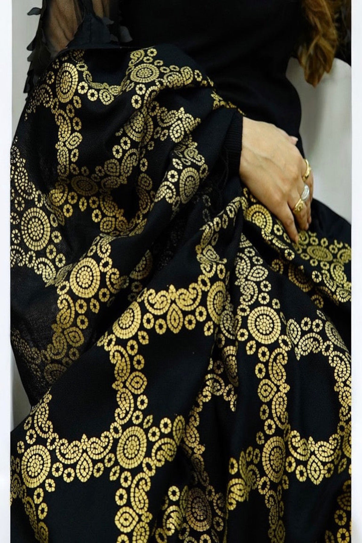 A woman sitting on a classic beige sofa, draped in a luxurious black shawl featuring an intricate, heavy golden circular jacquard pattern and traditional fringe details.