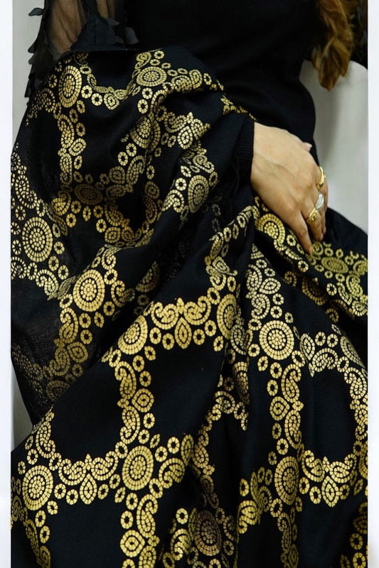 A woman sitting on a classic beige sofa, draped in a luxurious black shawl featuring an intricate, heavy golden circular jacquard pattern and traditional fringe details.