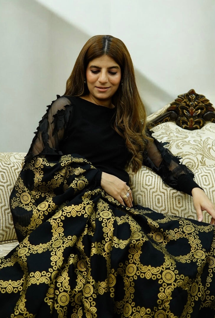A woman sitting on a classic beige sofa, draped in a luxurious black shawl featuring an intricate, heavy golden circular jacquard pattern and traditional fringe details.