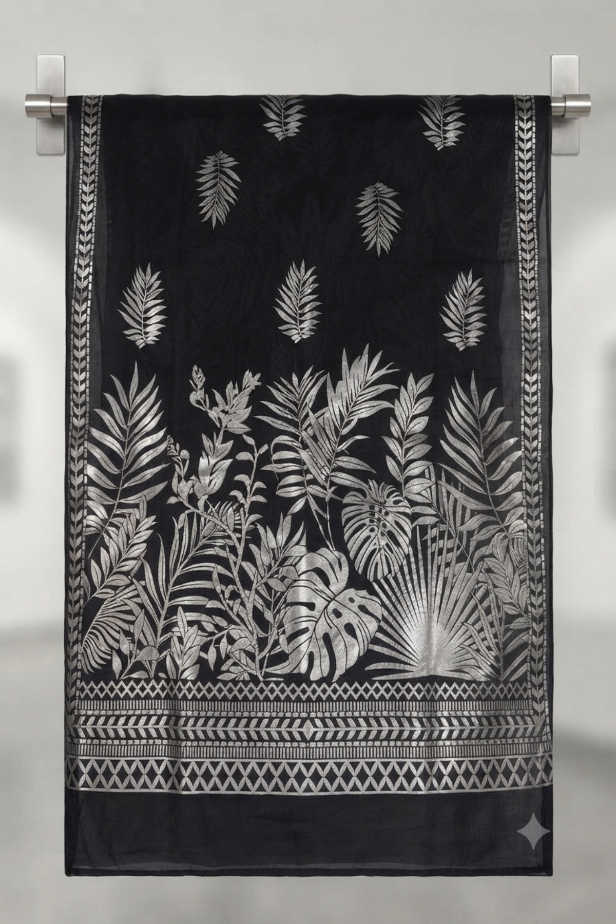 A black fabric dupatta, possibly organza or silk, hangs vertically from a brushed silver rod. It features a complex metallic silver foil print of various tropical leaves and foliage, including monstera, rising from a detailed geometric and laurel border at the bottom. The background is a softly blurred, modern gallery space
