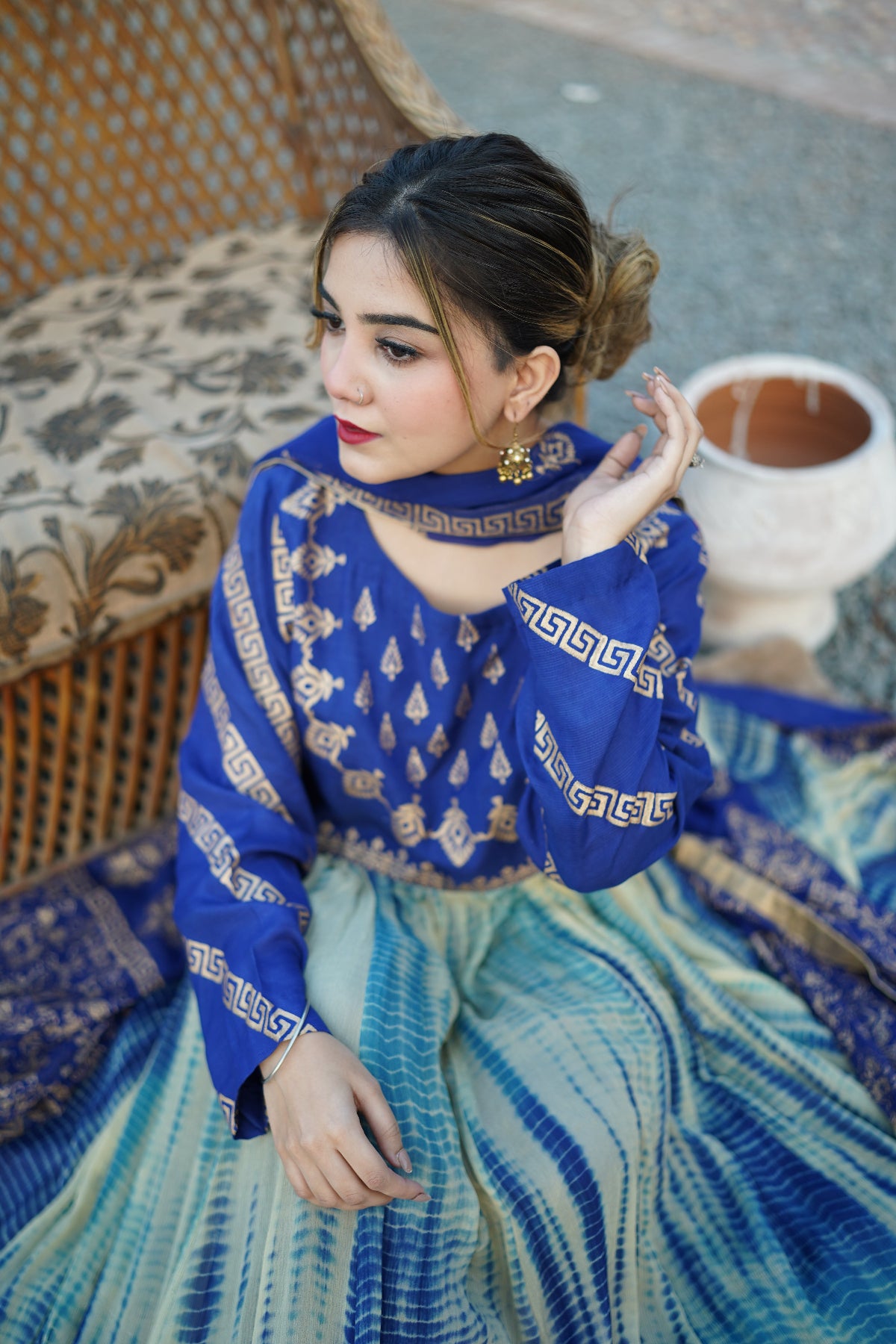 Woman in a blue and green traditional outfit sitting on a patterned chair with a cup of tea.