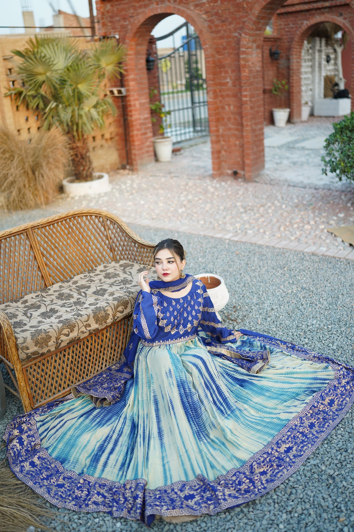 Woman in a blue and white traditional outfit sitting on a textured surface with a wicker chair and plants in the background.