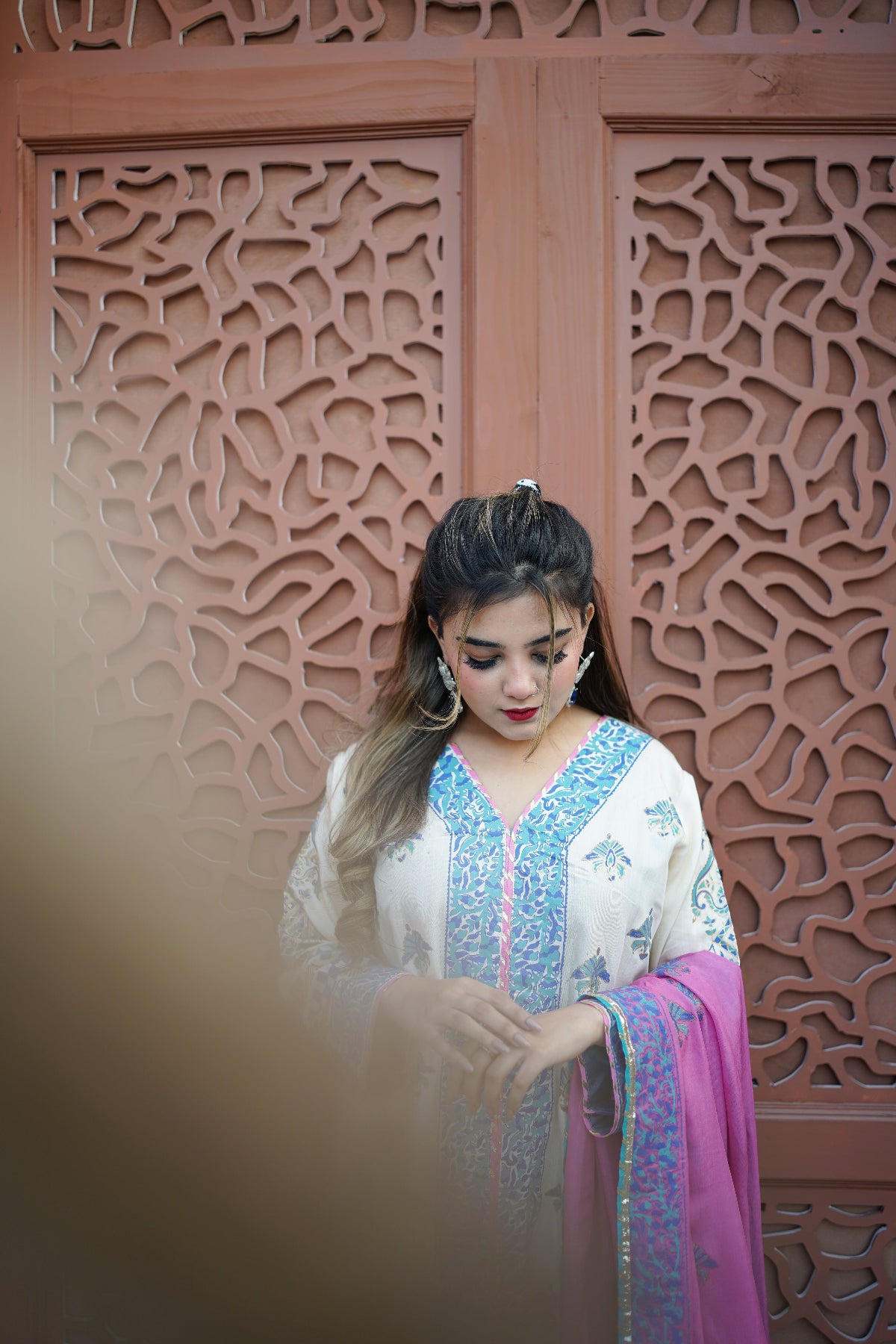 A woman wearing a traditional lemon and blue embroidered outfit with a pink dupatta, standing in front of a carved reddish-brown wooden screen.
