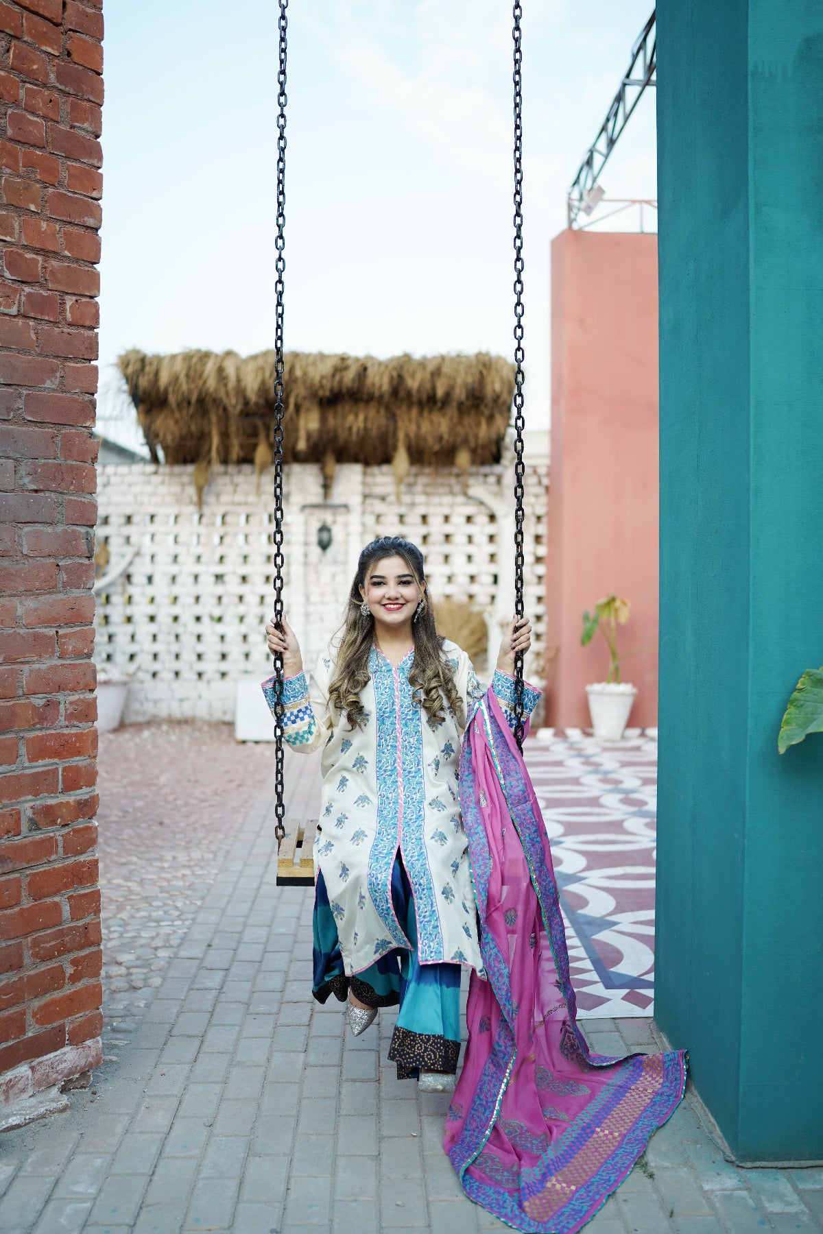 A woman wearing a traditional three-piece salwar kameez outfit in light blue and white with a vibrant pink and purple dupatta, sitting on a hanging swing in an outdoor courtyard.