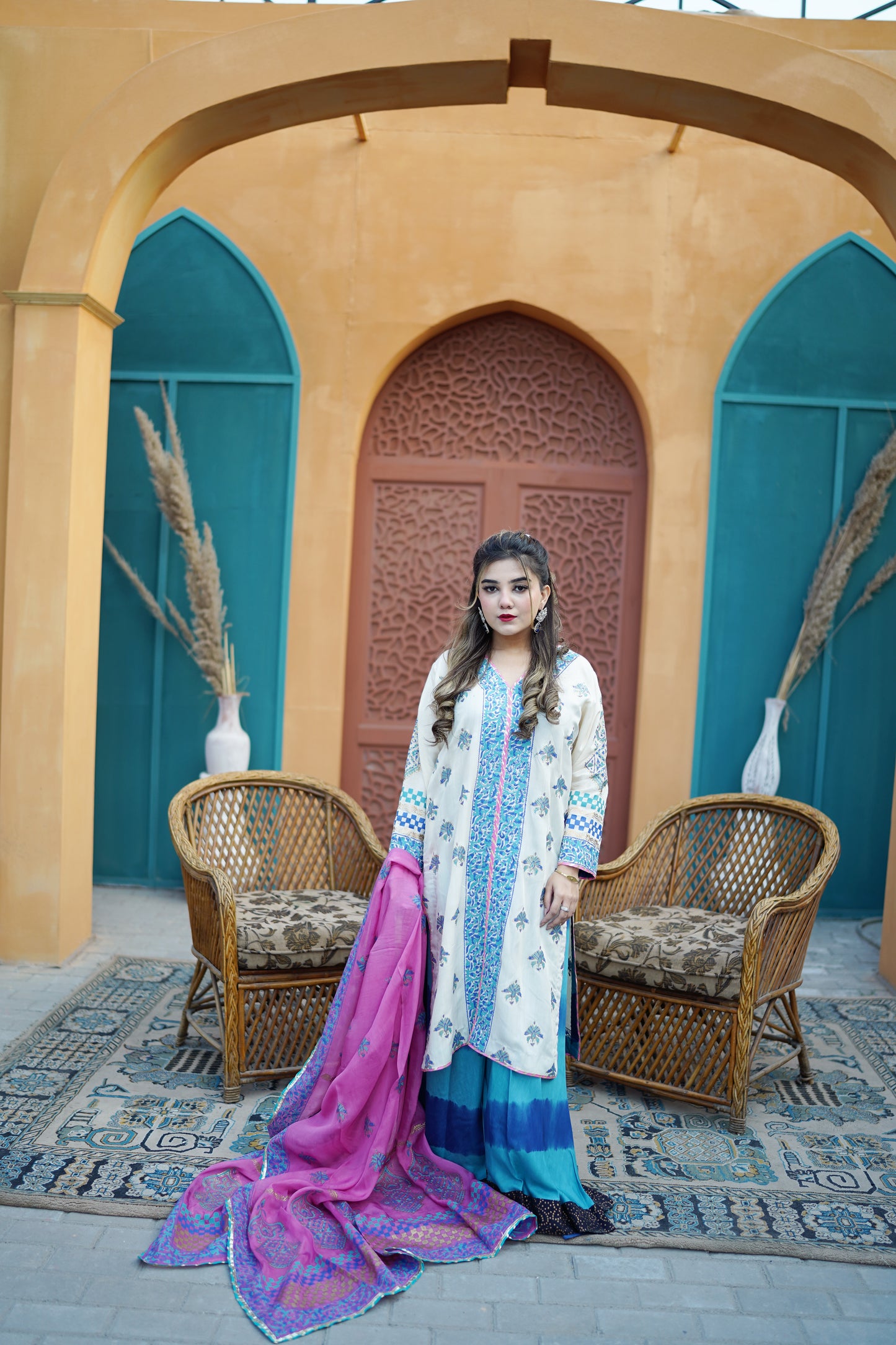 A woman stands in front of a traditional archway wearing a white kurta with blue floral embroidery, a blue and teal skirt, and a bright pink dupatta with purple borders. Two wicker chairs and a patterned rug are on the floor.