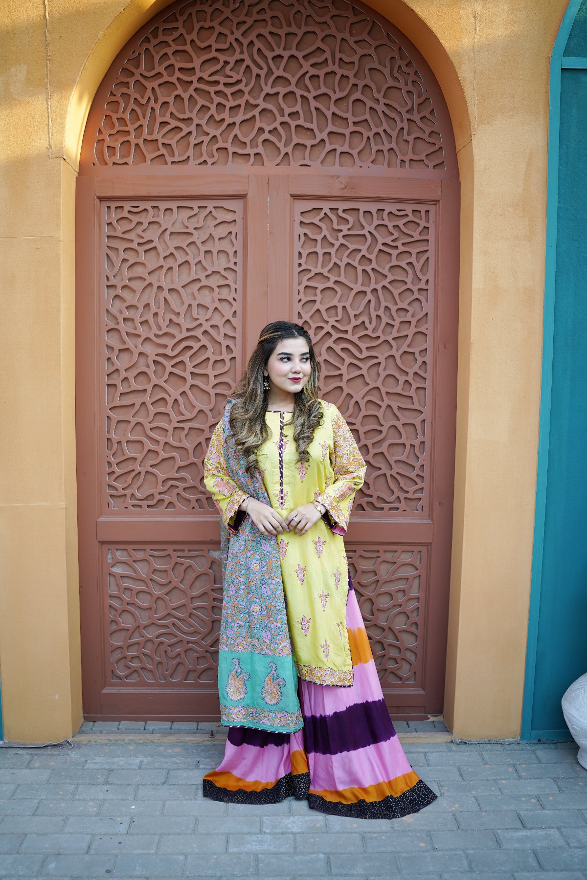 Model posing in a luxury 3-piece unstitched raw silk suit featuring a vibrant yellow shirt, a festive two-tone dyed palazzo with pink, purple, and orange horizontal stripes, and a patterned medium silk dupatta
