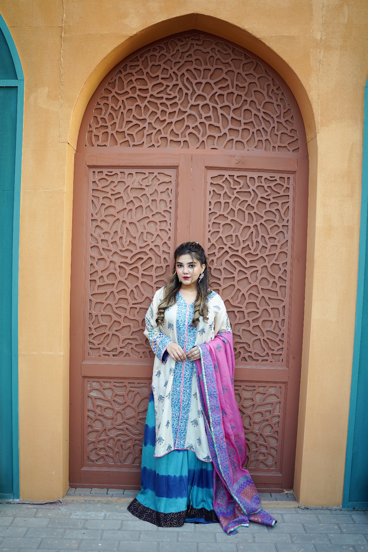 A woman stands in front of a large arched brown door with intricate geometric carvings, wearing a traditional outfit with a lemon block print  top, a blue and teal flared skirt, and a pink and purple dupatta.
