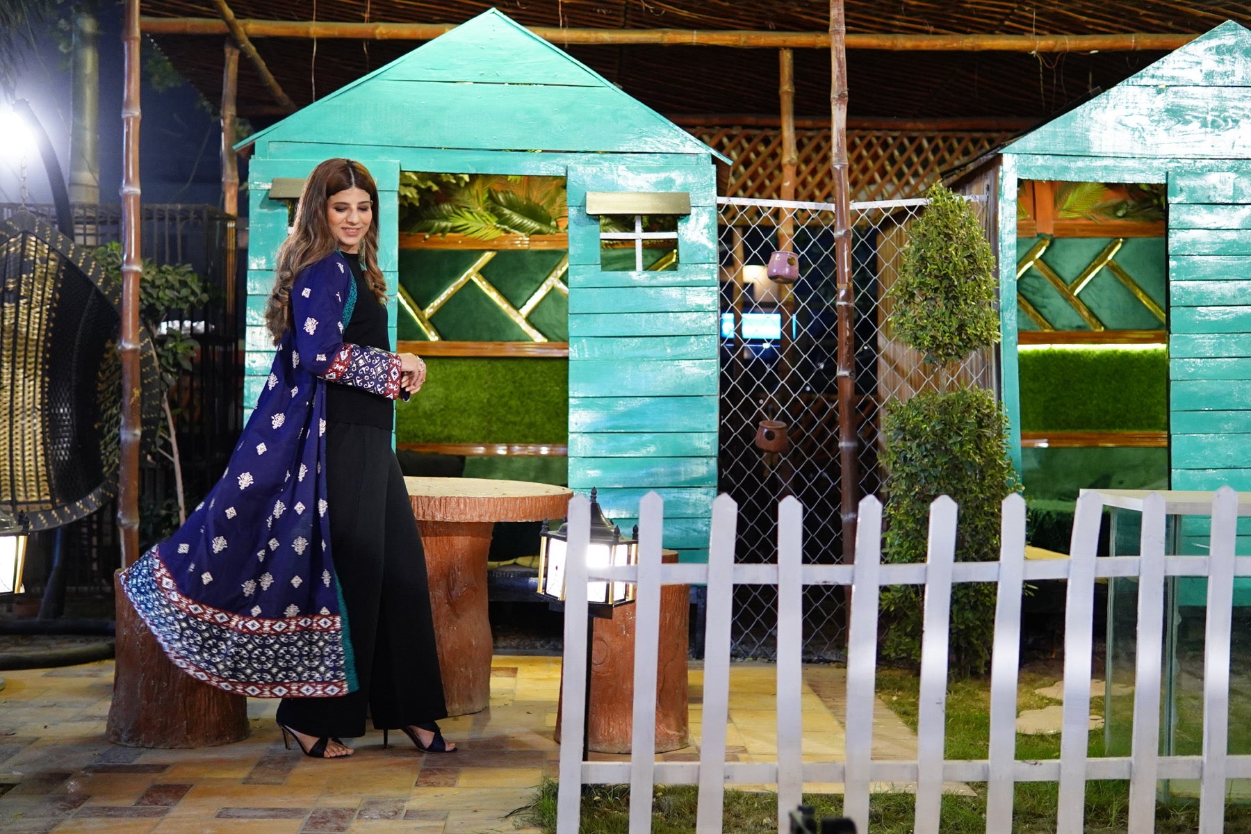 Woman in a blue block print gown standing in front of a small green wooden house with a white picket fence.