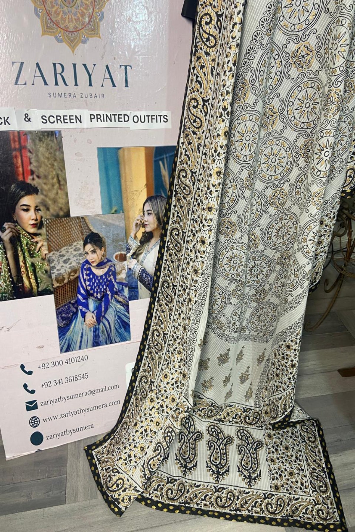 A folded and draped piece of Zariyat Sumera Zubair screen-printed ethnic wear fabric, showing intricate silver, black, and gold floral and paisley patterns on a light beige base. A promotional display card for 'Zariyat' is visible in the background.