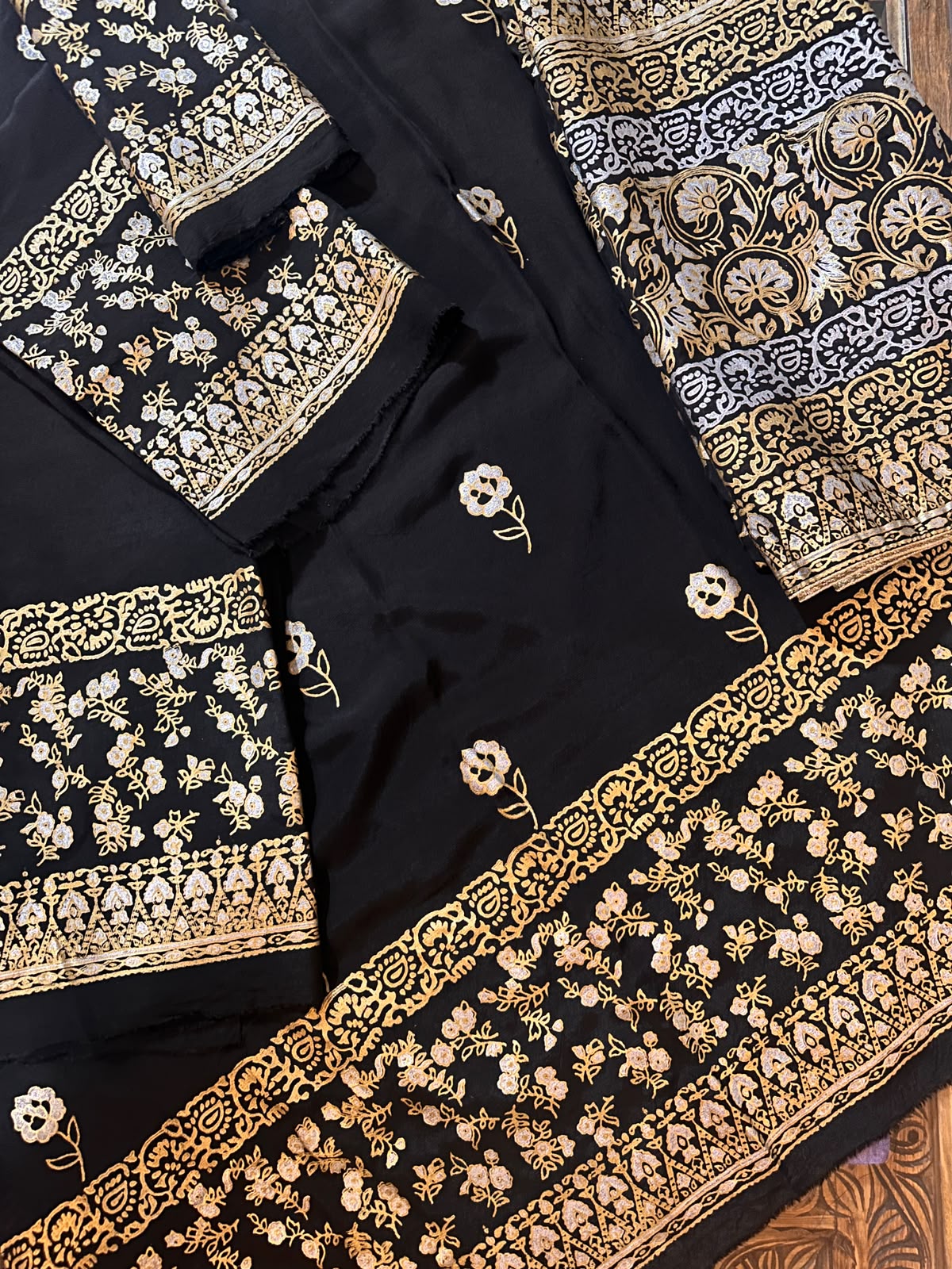 A close-up view of a Zariyat by Sumera 3-piece unstitched suit featuring a black fabric base with intricate gold floral block print patterns and traditional border designs