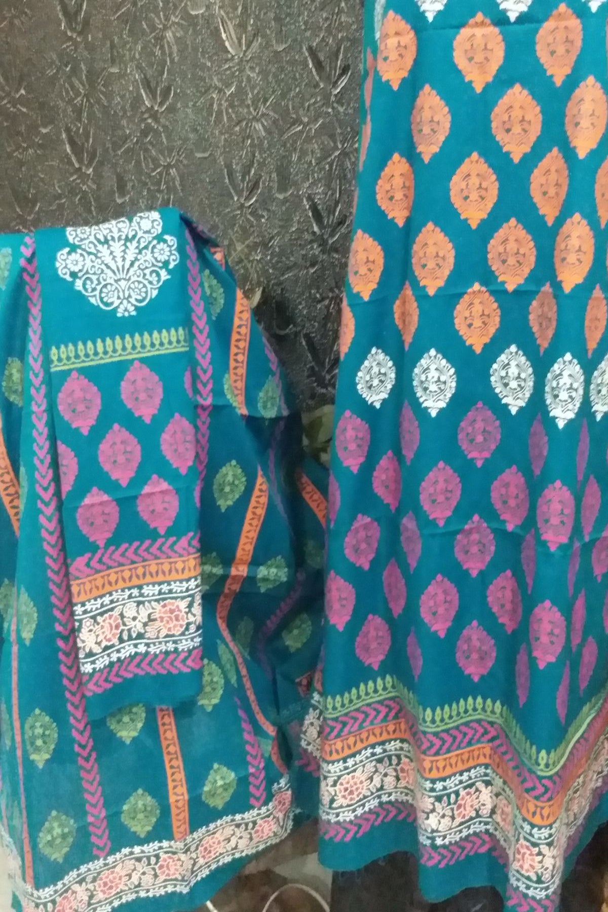A teal blue ethnic salwar suit set featuring a printed kurta with orange and white motifs,  matching fabric panel with colorful borders.